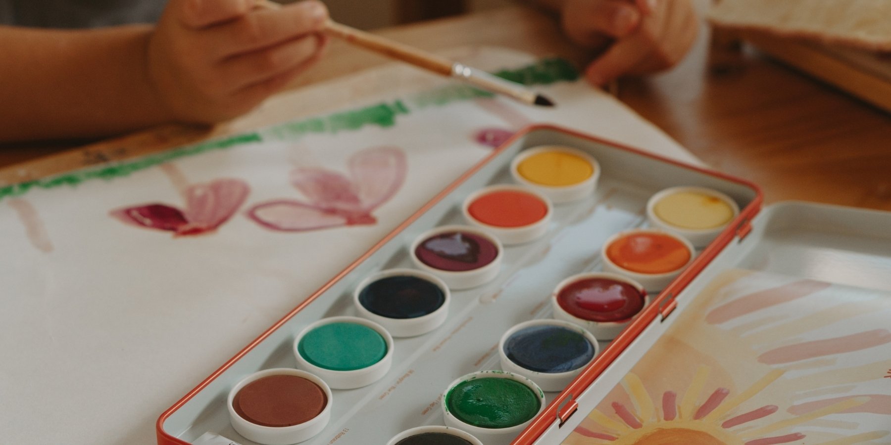 A close-up of a STOCKMAR watercolor paint palette with multiple coloured pans, placed on a wooden table next to a child's hands painting on white paper.