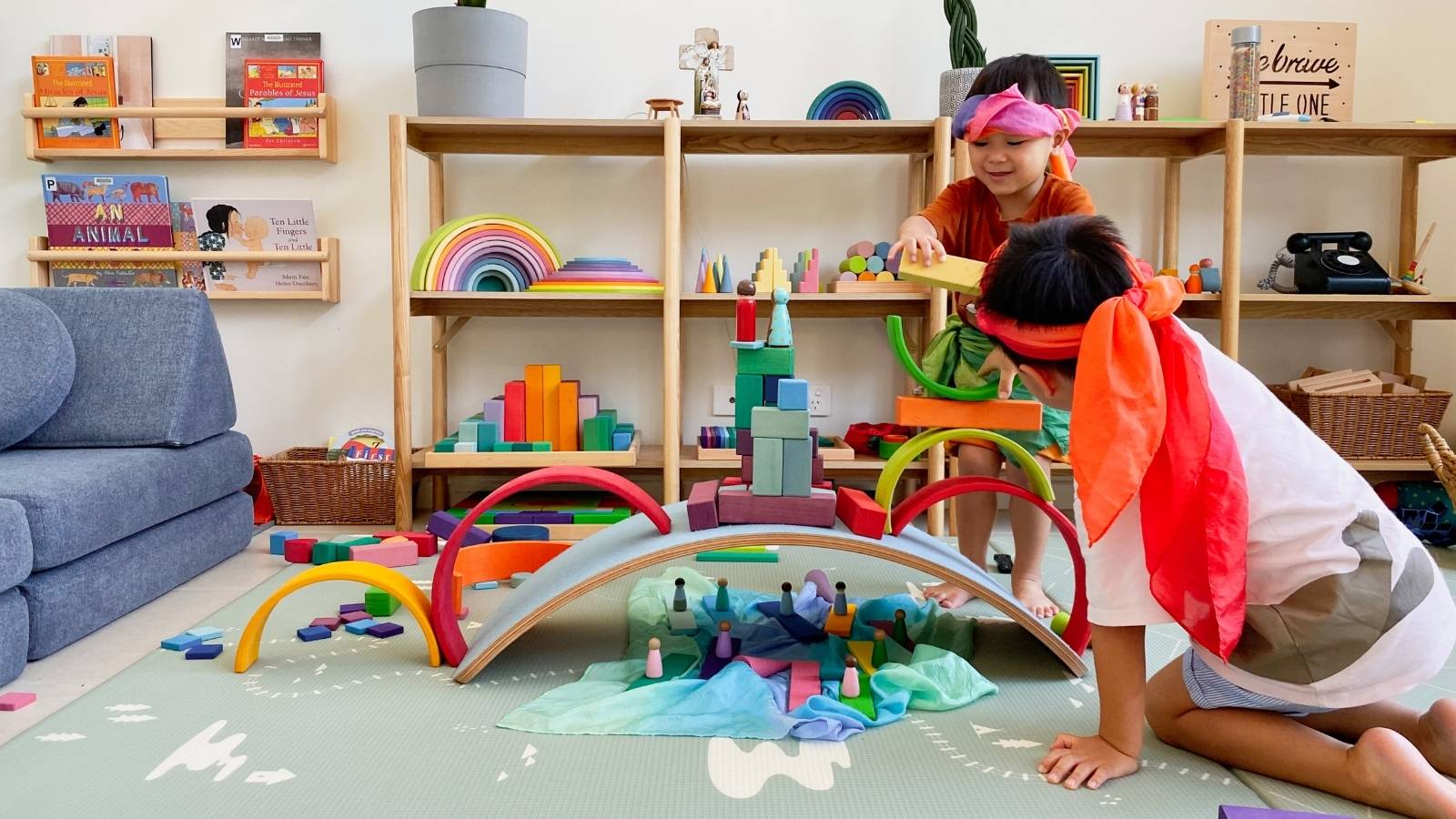 Two children playing with colourful wooden Grimm's rainbow arches and Sarah's Silks playsilks on a play mat in a bright children's room. Shelves in the background hold books, toys, and decor.