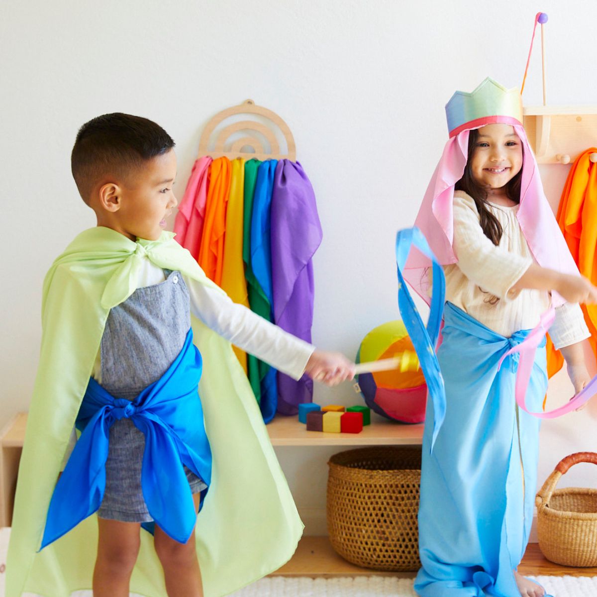 Two children playing dress-up with Sarah's Silks playsilks  in a bright indoor playroom. The boy on the left wears a pale yellow cape tied at the waist over a gray shirt and denim shorts.