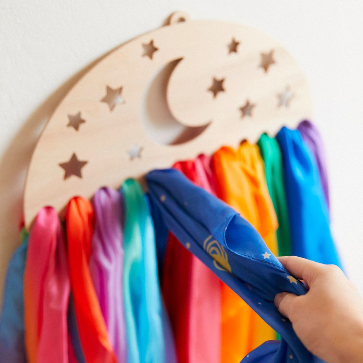 A wooden crescent moon and star wall display with colourful Sarah's Silks playsilks hanging from it, shown against a plain light wall. A hand is holding a playsilk with a small yellow moon and star print.