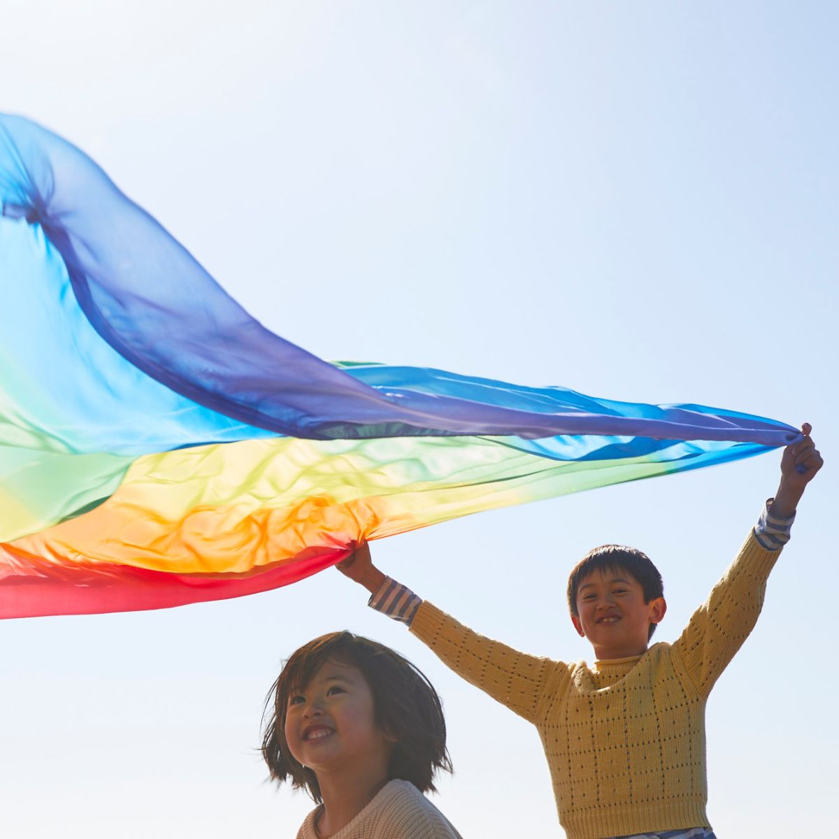 A child in a yellow sweater holds up a large rainbow-coloured Sarah's Silks playsilk outdoors under a clear blue sky, with another child partially visible below.