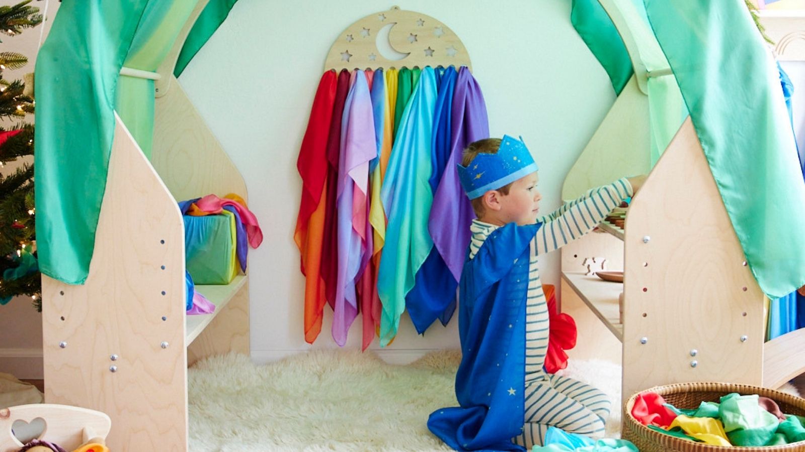 Child engaging in open-ended imaginative play in a wooden cubby draped with a giant Sarah's Silks playsilk and a selection of playsilks hanging up in the background