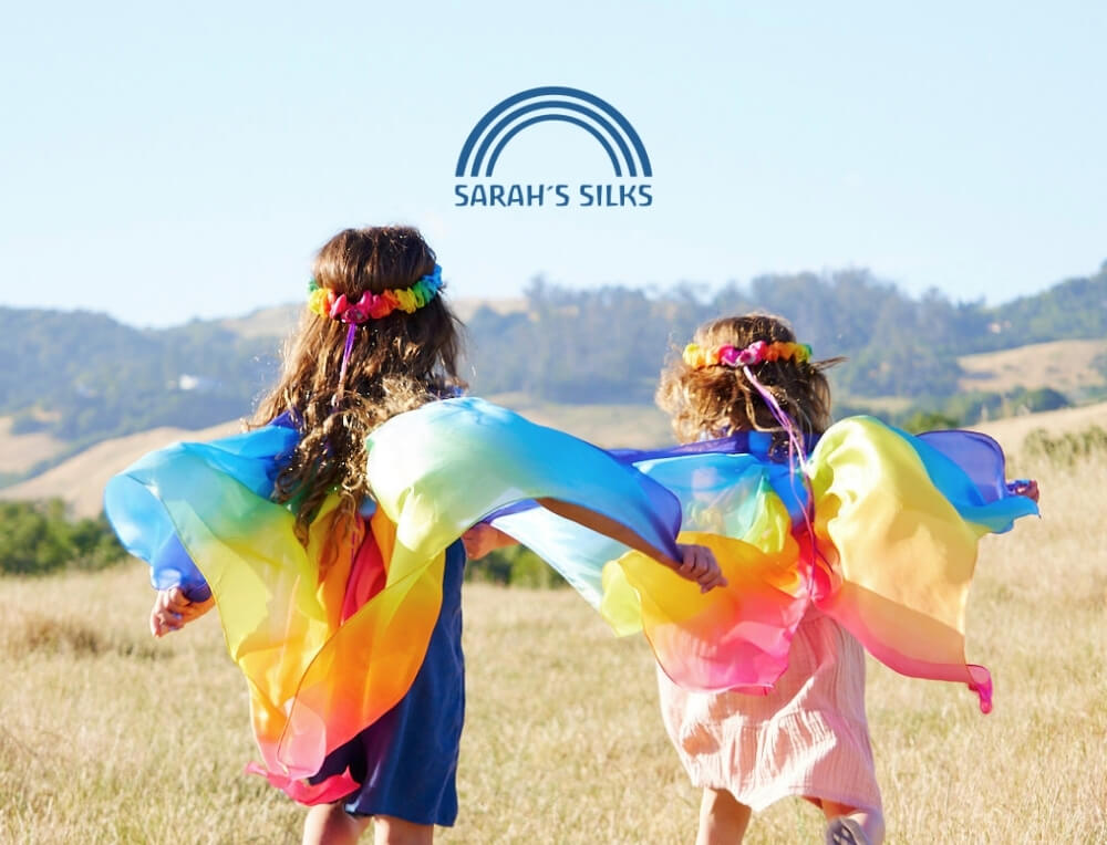 Sarah's Silks lifestyle image with logo, featuring two children seen from behind in a grassy field, wearing colourful rainbow silk wings and garland crowns – Oskar's Wooden Ark