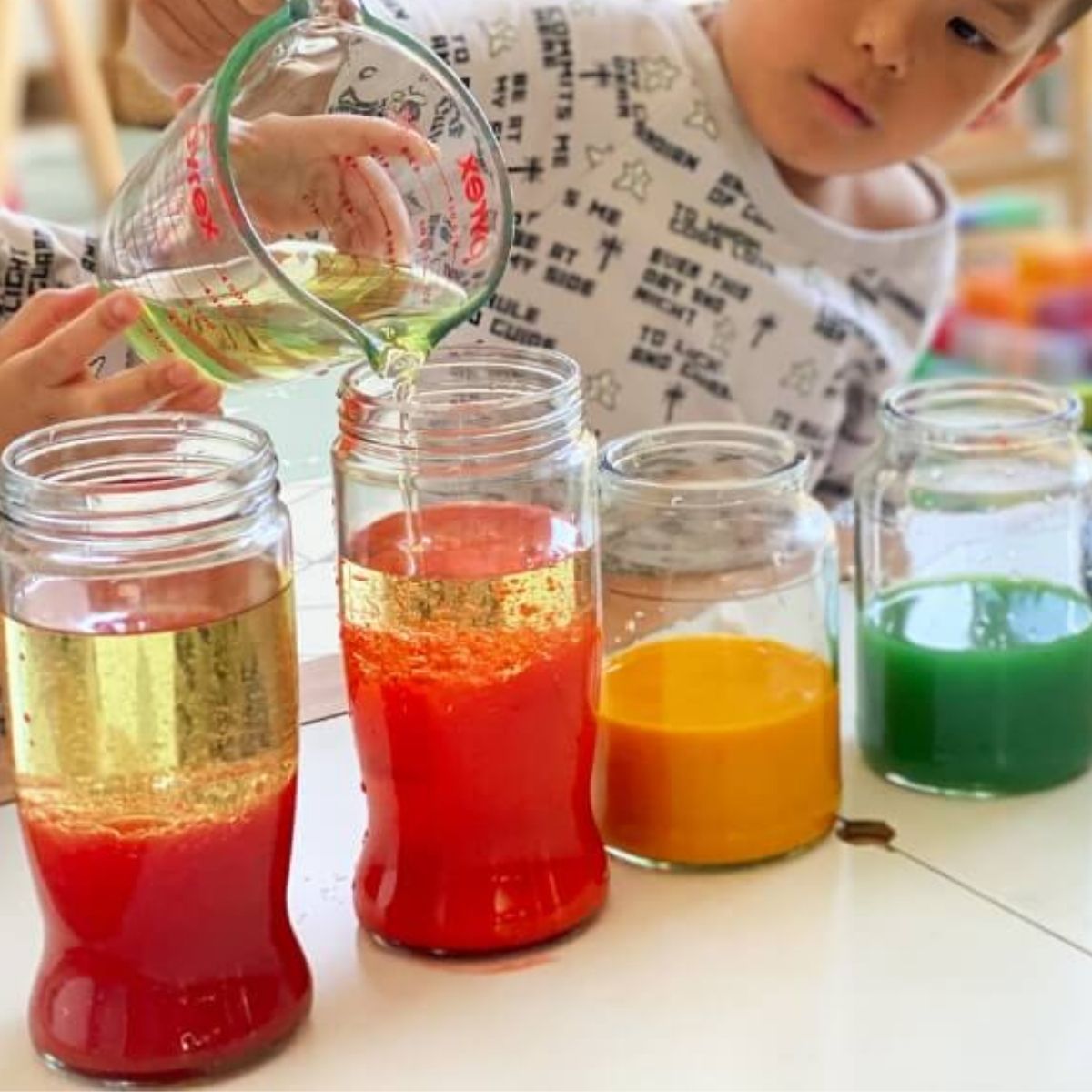 Child pouring in a glass jar filled with layered bright yellow, red, and green coloured water sitting on a table.