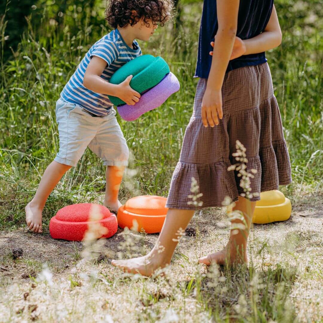 A child in a t-shirt and shorts holds a stack of colourful Stapelstein stepping stones outdoors, with another person standing nearby and several bright Stapelstein stones on the grassy ground.