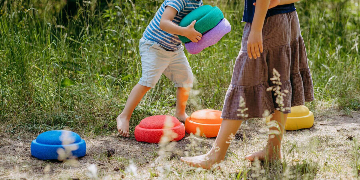 A child in a t-shirt and shorts holds a stack of colourful Stapelstein stepping stones outdoors, with another person standing nearby and several bright Stapelstein stones on the grassy ground.