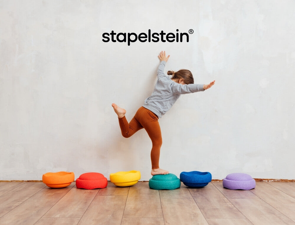 Stapelstein lifestyle image with logo, featuring a child balancing on colourful circular Stapelstein stepping stones arranged in a row against a textured off-white wall – Oskar's Wooden Ark