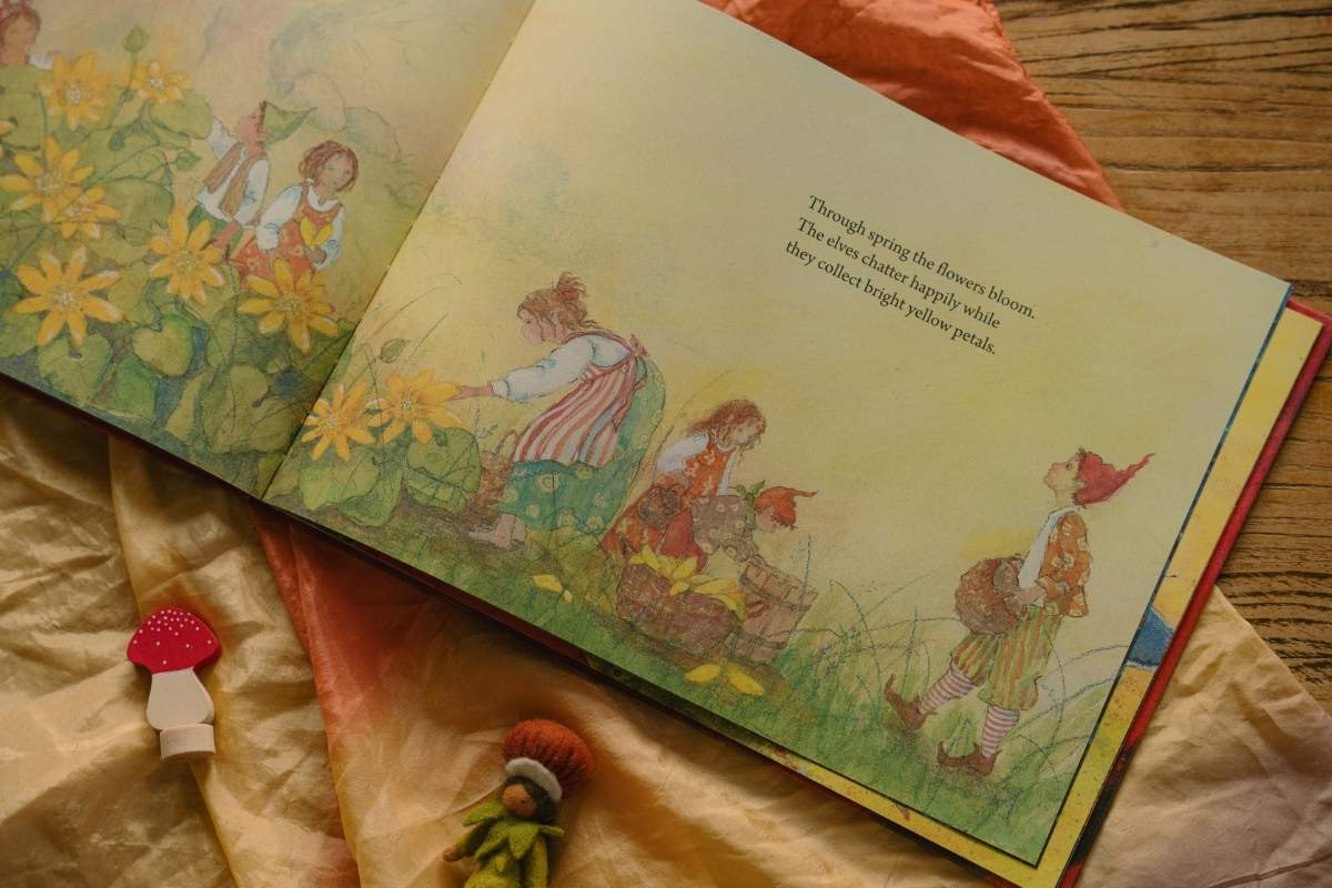 An open children's picture book spread on draped fabric, showing a colorful illustration of children in a flower field with text on the right page. Small mushroom and bird figurines sit nearby on the fabric.