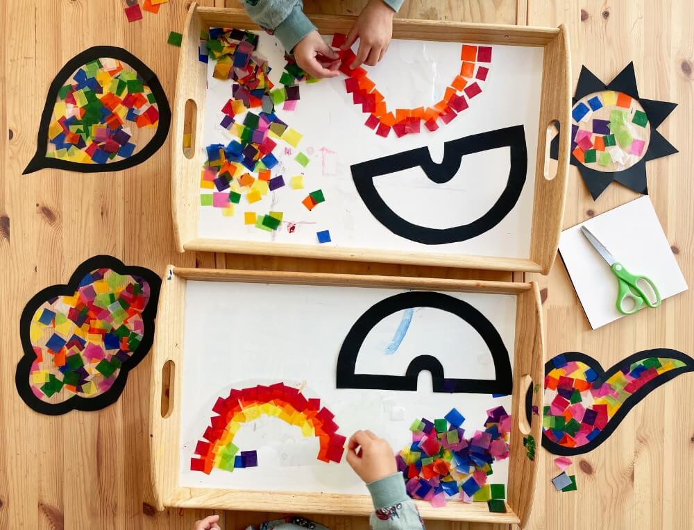 Child creating a mosaic collage with waxed kite paper squares on sticky contact shapes for a DIY sun catcher