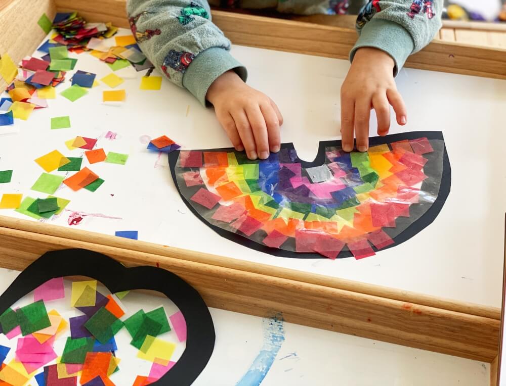A child making a sun catcher with colourful waxed kite paper squares on sticky contact shapes.