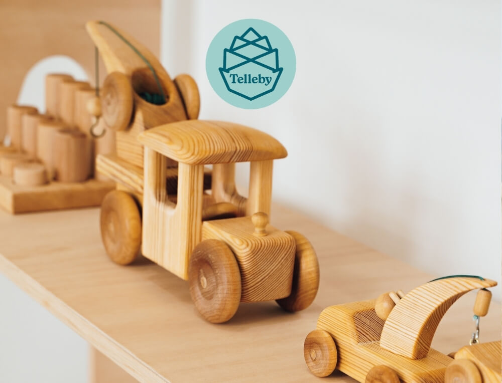 Telleby Debresk lifestyle image with logo, featuring handcrafted wooden toy vehicles on a light wood shelf, featuring a central wooden truck and a smaller car in the foreground – Oskar's Wooden Ark