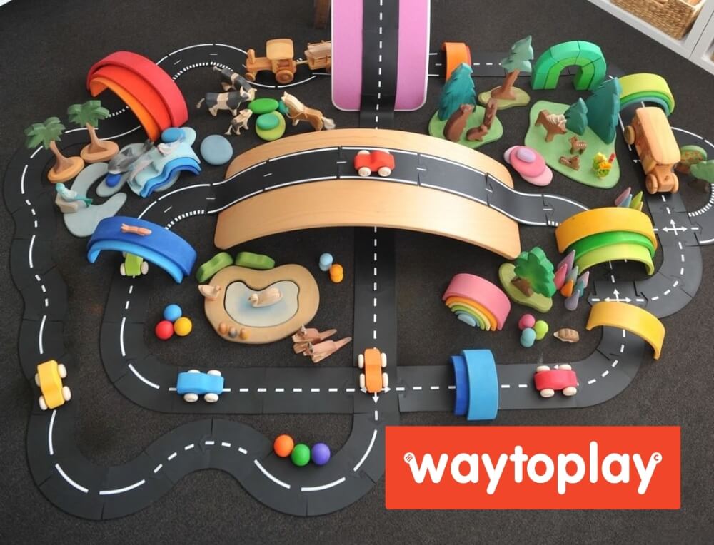 A WaytoPlay road playset with colourful toy cars, bridges, tunnels, trees, and a winding black road on a dark surface. A red WaytoPlay logo label is visible in the bottom right corner.
