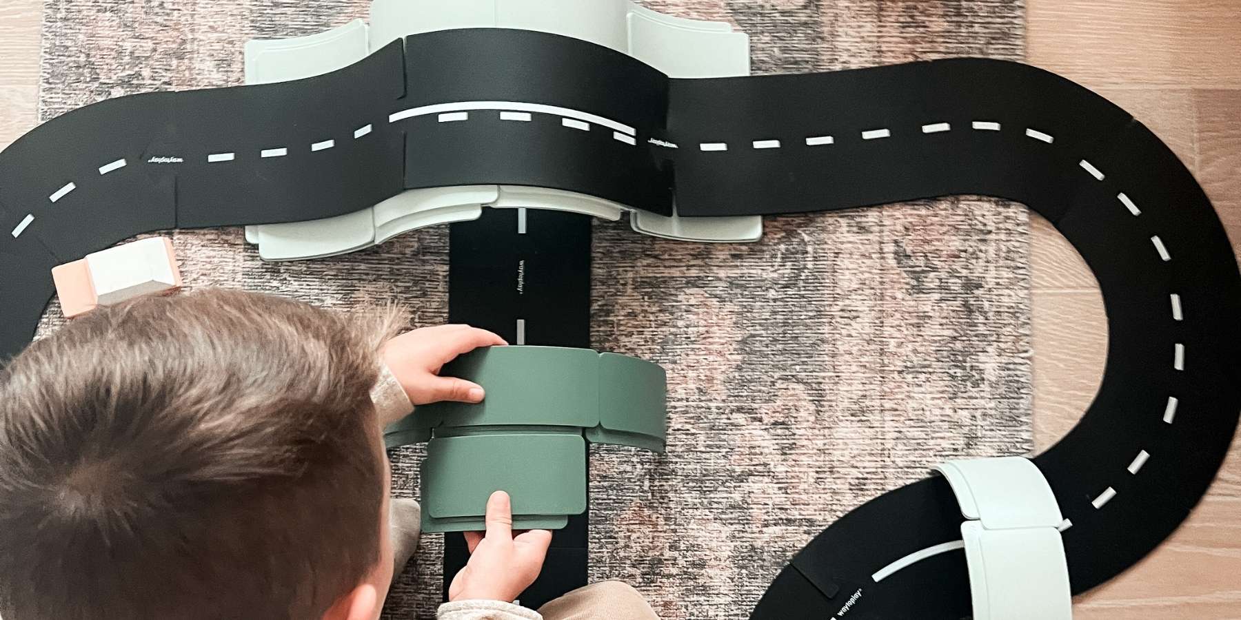A child's hands and head are visible as they play with a WaytoPlay toy road track and arches set on a textured rug. 