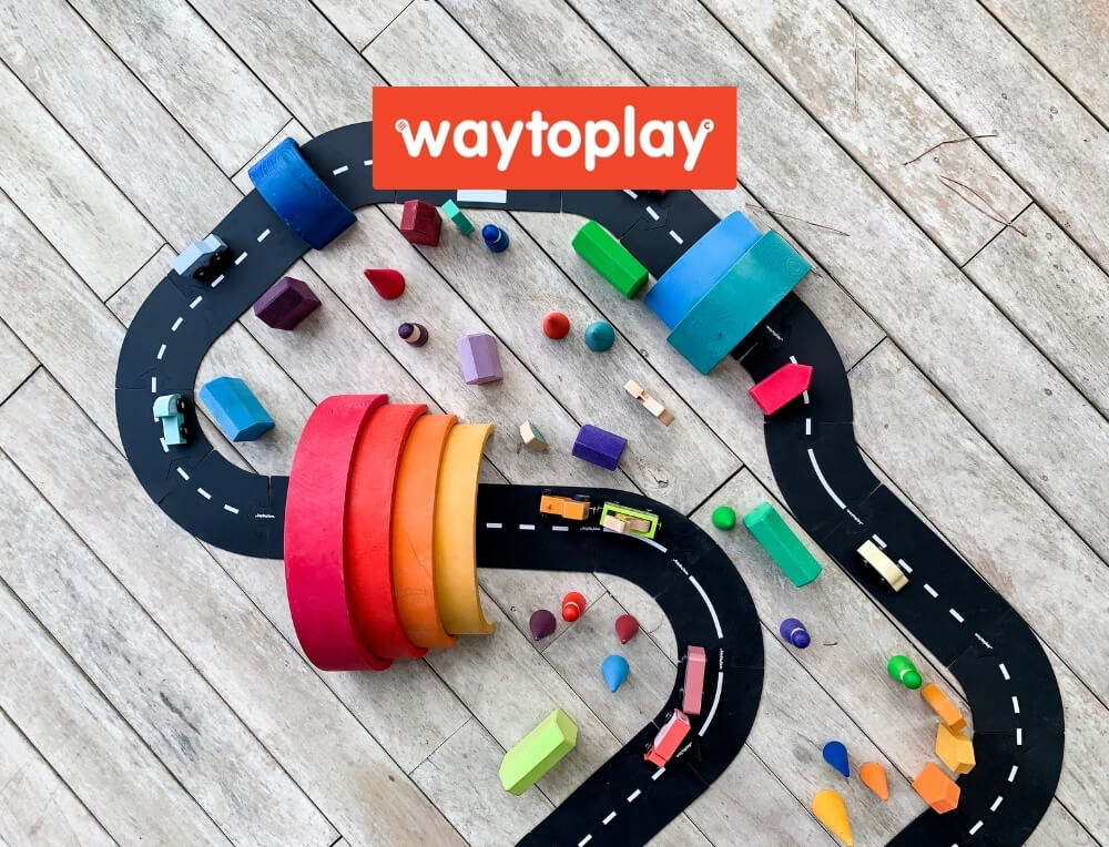 Waytoplay Roads lifestyle image with logo, featuring a winding black road with white dashed lines, rainbow-coloured tunnel pieces, and small multicoloured toy cars  – Oskar's Wooden Ark