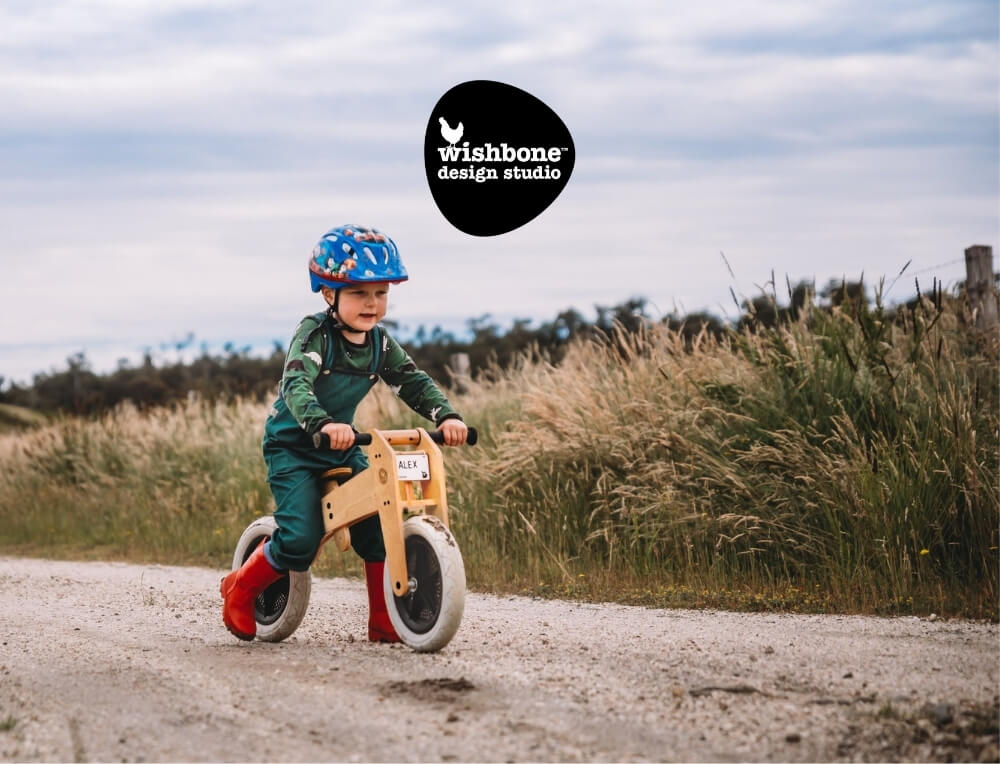 Wishbone Design Studio lifestyle image with logo, featuring a child riding a wooden balance bike outdoors on a gravel path – Oskar's Wooden Ark