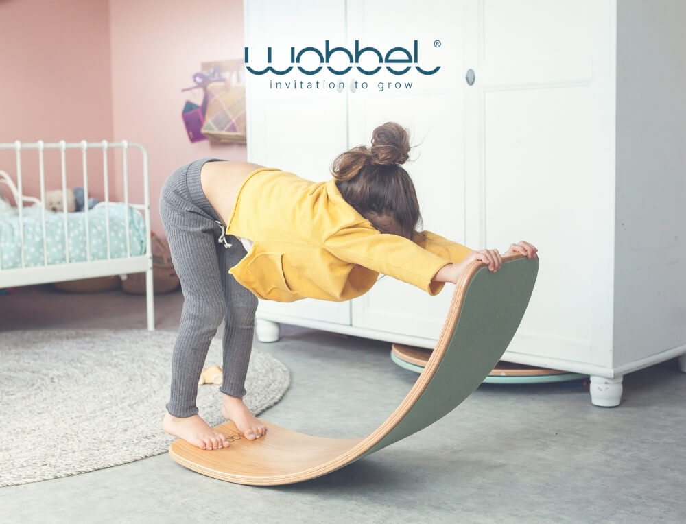 Wobbel Board lifestyle image with logo, featuring a child balancing on a Wobbel brand curved wooden balance board in a child's bedroom – Oskar's Wooden Ark