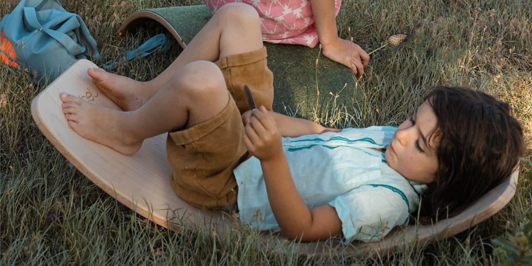 A child lying on a wooden Wobbel board outdoors in tall grass, wearing a light blue shirt and brown shorts, with another child sitting on a Wobbel board in the background.