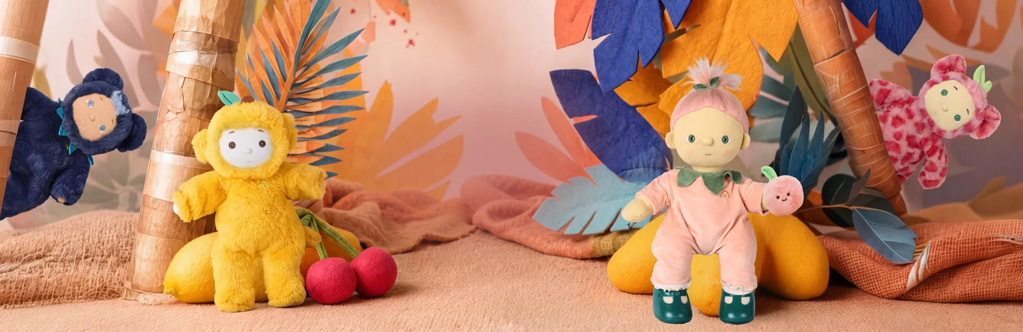 A collection of colorful Dinkum Doll  toys styled in a tropical-themed setting with palm leaves, beach umbrellas, and a sandy background. The toys include a yellow character with red cherries, a pink character with green shoes, and other partially visible plush figures.