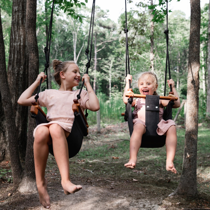 A little girl laughing while swinging outdoors in the woods using SOLVEJ Convertible Baby & Toddler Swing with an older girl swinging on a SOLVEJ Child Swing