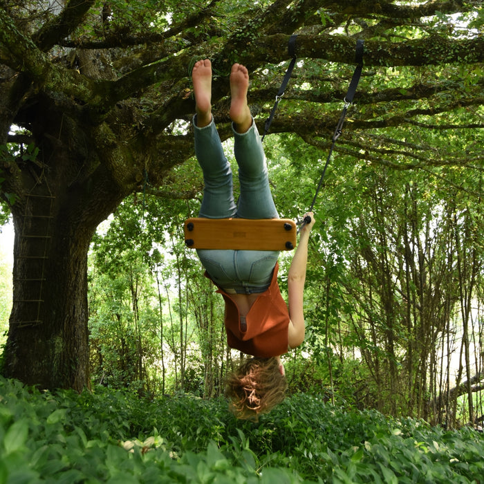 A woman doing a play swing flip trick in the woods using the SOLVEJ Traditional Wooden Board Swing with Black Rope hung from a tree