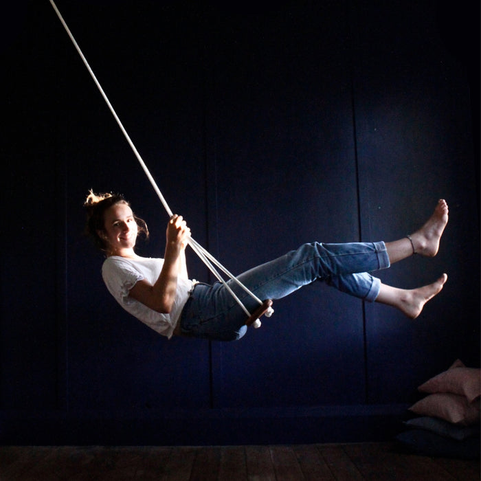 A woman smiling while swinging on the SOLVEJ Wooden Traditional Board Swing with White Rope hung indoors