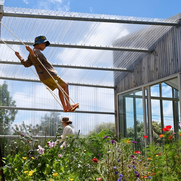 A boy in a greenhouse doing a play swing trick while standing on the the  SOLVEJ Wooden Traditional Board Swing with White Ropes from Australia